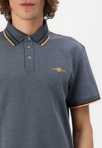 Mørkegrå polo skjorte med en struktureret stof. Har en kontrasterende krave med en gul og marineblå stribe, broderet logo med "1962."