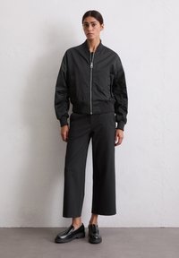 Woman standing against a white wall wearing a black zip-up bomber jacket, wide-leg cropped black pants, and black loafers.