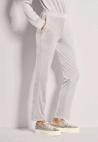 Persoon in lichtgrijze velours broek en sneakers met slangenhuidpatroon, staand met één hand in de zak tegen een effen achtergrond.