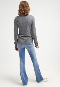 Femme aux cheveux bruns bouclés portant un t-shirt gris à manches longues, un jean évasé bleu clair et des baskets beiges, debout devant un fond blanc uni.