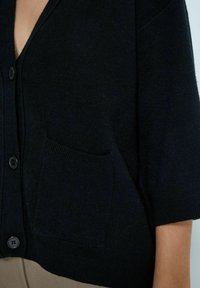 Cardigan noir avec un col en V, des manches courtes et une poche avant unique. Fabriqué en tissu doux avec un détail texturé et une fermeture à boutons.