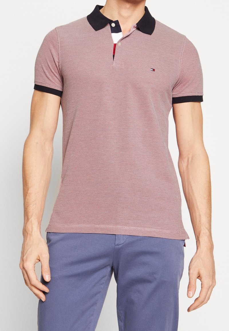 Homme portant un polo mauve clair avec un col et des bords de manches noirs, associé à un pantalon bleu, debout devant un fond uni.