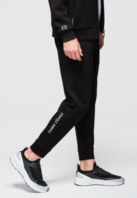 Personne portant un pantalon de jogging noir avec le texte « OMBRE EXIMIO » sur la jambe et des chaussures de sport noires et blanches, debout devant un fond blanc.