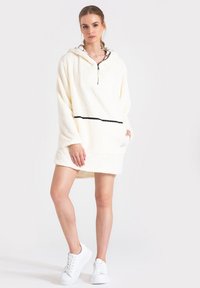 Kremowa, polarowa sukienka z kapturem, z półzamkiem, przednią kieszenią, czarnym paskiem i wzorzystym wykończeniem kaptura. Noszona z białymi trampkami.