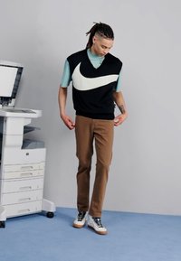 Schwarzer ärmelloser Strickpullover mit großem weißen Nike-Logo, getragen über einem hellblauen T-Shirt, kombiniert mit braunen Hosen und weißen High-Top-Sneakern.