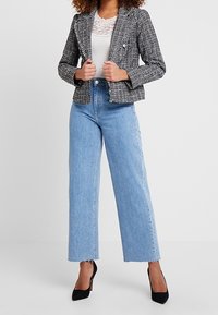 Kvinna klädd i en svart och vit tweedjacka, vit spetsblus, högmidjade vida blå jeans och svarta spetsiga klackskor.