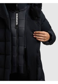 Giacca a vento nera con texture trapuntata, dotata di una patta con zip, cappuccio regolabile e un grande tag del brand sulla parte anteriore.