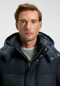 Marinblå puffjacka med hög krage, silverfärgade detaljer, sydda paneler, bärs över en grå tröja, med dragkedja fram och tryckknappsdetaljer.