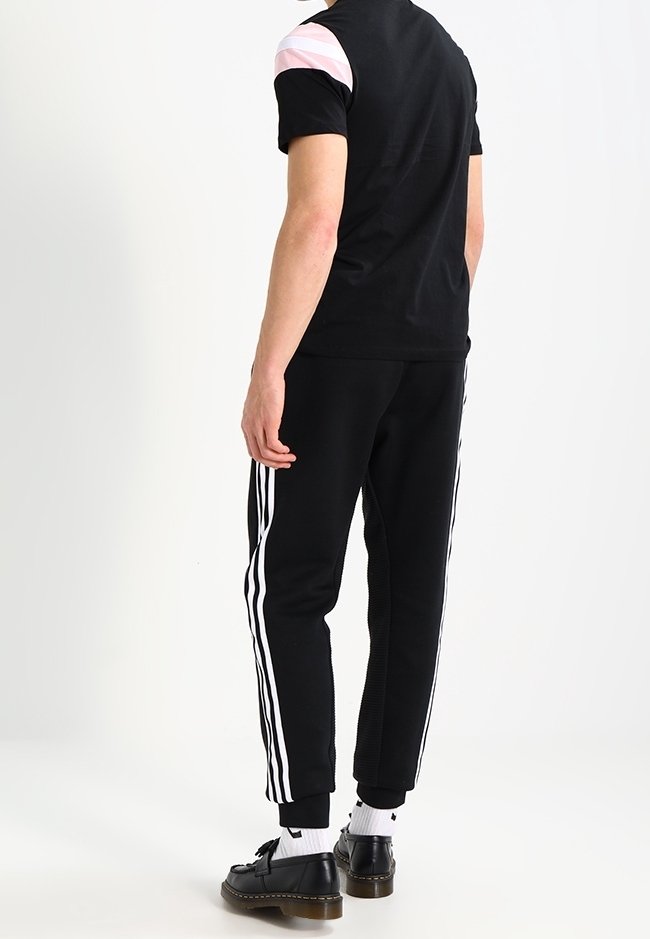 Uomo che indossa pantaloni da ginnastica neri con strisce laterali bianche, t-shirt nera con dettagli rosa e bianchi sulle spalle, calzini bianchi e scarpe nere, di spalle.