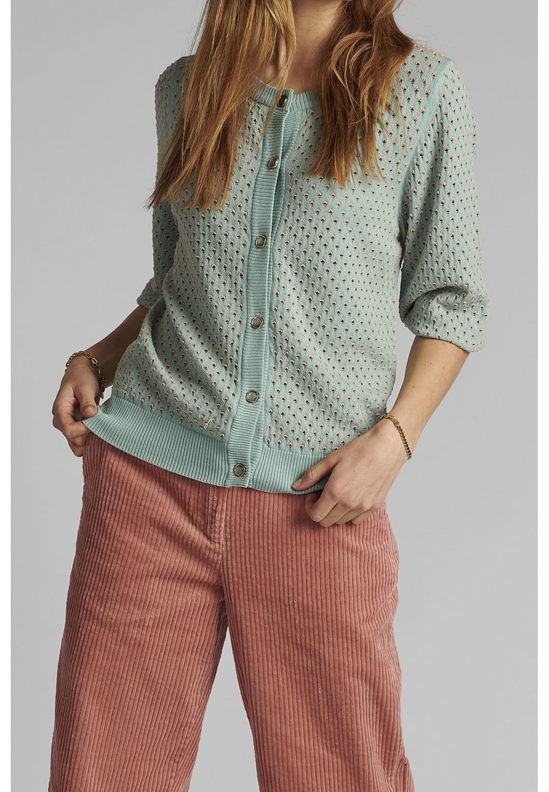 Femme portant un cardigan boutonné bleu clair à motif avec des manches trois quarts et un pantalon en velours côtelé rose poussiéreux, tenant l'ourlet du cardigan.
