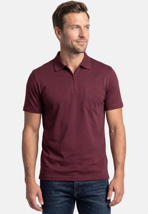 Mężczyzna ubrany w bordową koszulkę polo z krótkim rękawem, z suwakiem z przodu i kieszenią na piersi, w połączeniu z ciemnoniebieskimi dżinsami.