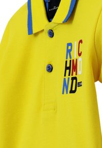Žluté polo tričko s modrým límečkem, s logem se znaky v červené, modré a černé. Dvě knoflíky na zapínání a strukturovaná tkanina.