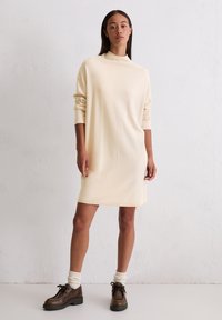 Gebreide jurk in licht crème, met een hoge hals en lange mouwen. Gecombineerd met donkerbruine schoenen en crèmekleurige sokken. Glad textuur.