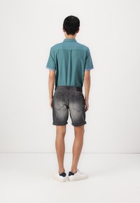 T-shirt en teal à manches courtes avec des accents bleus, associé à un short en denim gris. Le short est retroussé au niveau de l'ourlet, porté avec des baskets blanches.