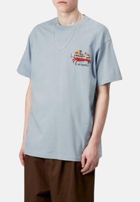 Camiseta de algodón azul claro con mangas cortas, que presenta un logo gráfico y un diseño de cangrejo en el lado izquierdo del pecho. Corte relajado, escote redondo.