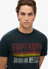 Sort bomulds t-shirt med "SUPERDRY" tekst i rød og grå samt vandrette striber i rød, gul og sort. Rund halsudskæring, korte ærmer.