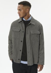 Mand iført en sort og beige ternet jakke over en sort sweater og en blåstribet skjorte, som ser til sin venstre side med hænderne i lommerne.