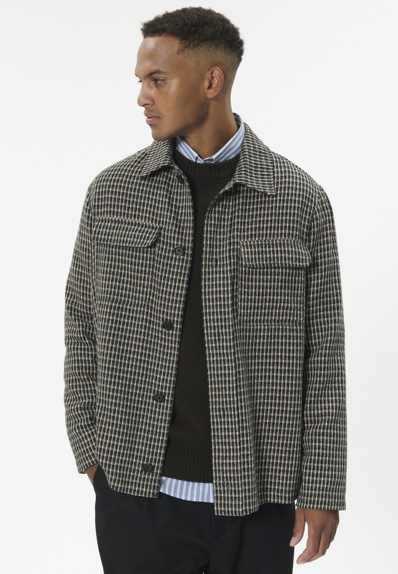 Mand iført en sort og beige ternet jakke over en sort sweater og en blåstribet skjorte, som ser til sin venstre side med hænderne i lommerne.