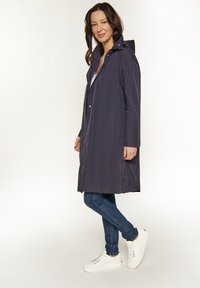 Impermeabile blu navy con cappuccio, maniche lunghe, bottoni a pressione, due tasche laterali e una vestibilità comoda, abbinato a jeans blu e sneakers bianche.
