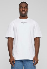 Biela bavlnená tričko s okrúhlym výstrihom, krátkymi rukávmi a čiernou grafickou potlačou podpisu na hrudi. Uvoľnený strih, ľahký materiál.