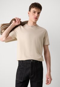 T-shirt beige à manches courtes avec un col rond et un petit logo "BOSS" au col, associé à un jean en denim foncé. Design décontracté.