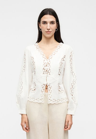 Femme aux cheveux longs et foncés portant un chemisier blanc à manches longues avec broderie florale découpée et un pantalon beige, debout devant un fond uni.