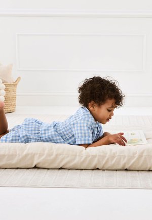 Vastsündinu sinist ruudulises riietuses lebamas patjal, lugemas väikest raamatut heledal vaibal heledas toas.