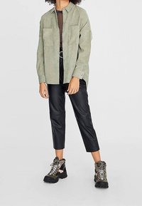 Persona che indossa una camicia button-up oversize di un verde chiaro, pantaloni neri a 3/4 e scarpe da trekking verdi con suole spesse e lacci.