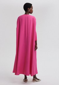 Flödande rosa kappa klänning, långa ärmar, texturerad tyg, A-linjeformad silhuett, ankellång, stängning i nacken bak, bärs med ljusa skor.
