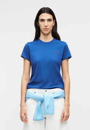 Γυναίκα με μακριά σκούρα μαλλιά φορά μπλε κοντομάνικη μπλούζα και λευκό παντελόνι, με ένα ανοιχτό μπλε πουλόβερ δεμένο γύρω από τη μέση.