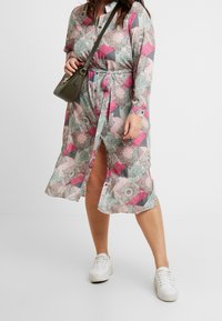 Robe à motifs floraux dans des teintes de rose, bleu et gris, avec une taille nouée, un devant boutonné et de longues manches. Associée à des baskets blanches.