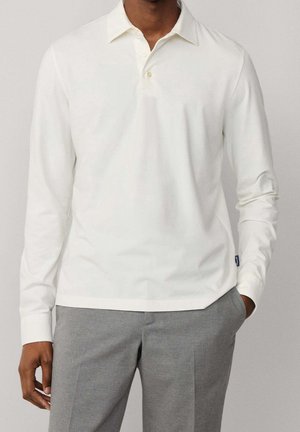 Homme portant un polo blanc uni à manches longues et un pantalon gris, debout devant un fond uni.