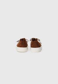 Une paire de baskets en suede marron avec des semelles en caoutchouc blanches, montrée de dos sur un fond blanc.
