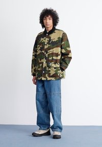 Καμουφλαρισμένο μπουφάν με κουμπιά μπροστά, μαύρο γιακά, δύο τσέπες και ετικέτα στο κάτω αριστερό μέρος; συνδυασμένο με μπλε φαρδιά τζιν παντελόνια και αθλητικά παπούτσια.