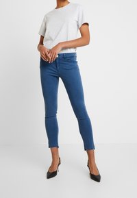 Person, der ein hellgraues kurzärmliges Hemd, enge blaue Knöchelhosen und schwarze hochhackige Slingbacks trägt, steht vor einem weißen Hintergrund.