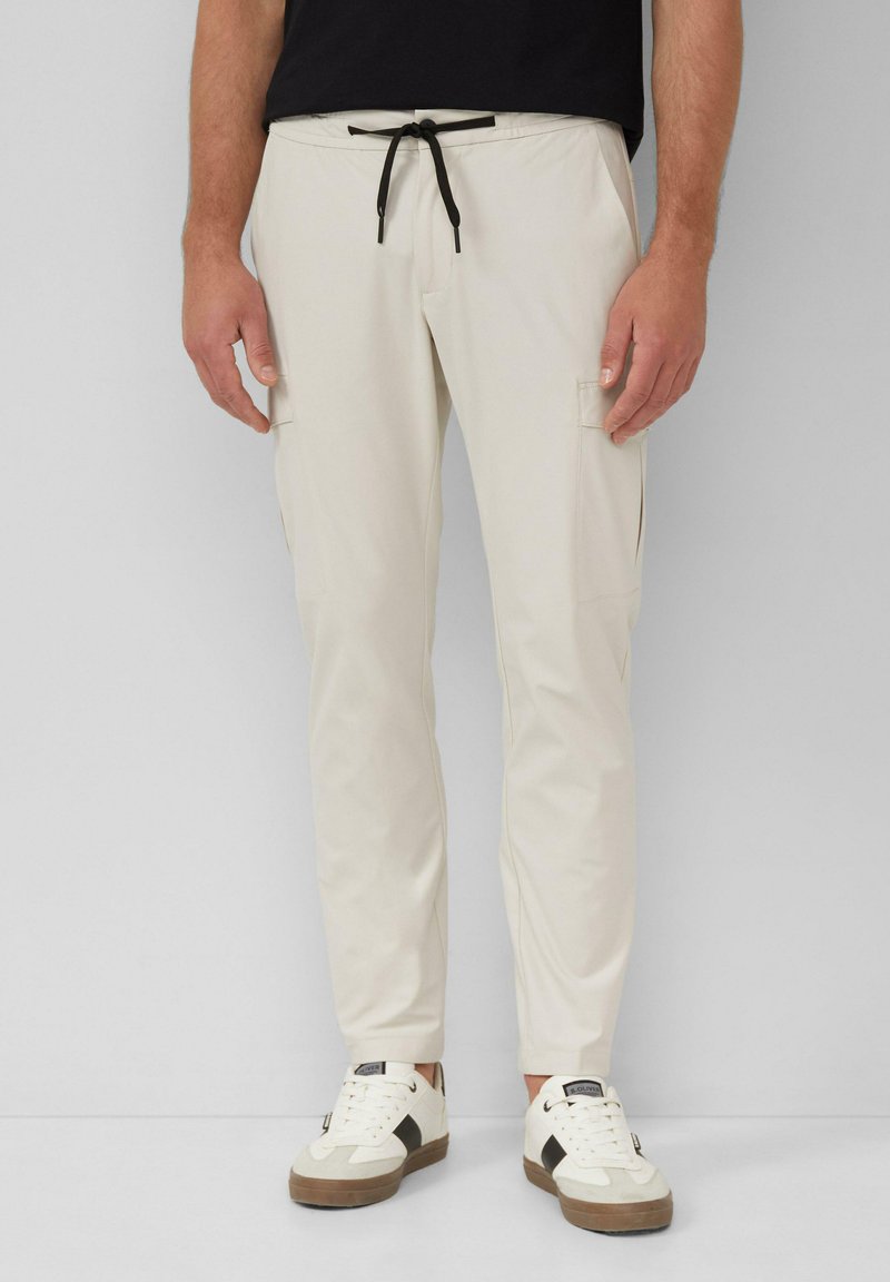 Lichtbeige cargo-broek met zwarte trekkoord, tapered fit, twee zijzakken en een gladde textuur, gecombineerd met witte sneakers.
