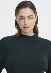 Pulover tricotat verde închis, cu guler înalt și mâneci lungi. Textură moale, croială lejeră, suprafață netedă, fără modele sau detalii accentuate.