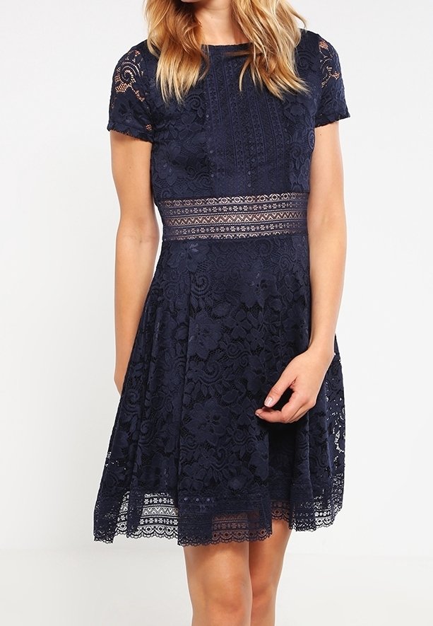 Femme portant une robe en dentelle bleu marine longueur genou, avec des manches courtes et une ceinture transparente à motif.