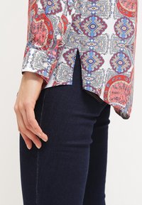 Langarmhemd mit einem bunten Paisleymuster in Rot, Blau und Weiß, ausgestattet mit einem Knopfverschluss an den Bündchen und einem gespaltenen Saum, kombiniert mit dunklen Jeans.