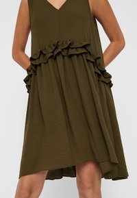 Vihreä oliivinvärinen hihaton mekko, jossa on V-pääntie, vyötäröllä rypytettyjä yksityiskohtia ja valuvat saumat, jotka kapenevat kohti helmaa.