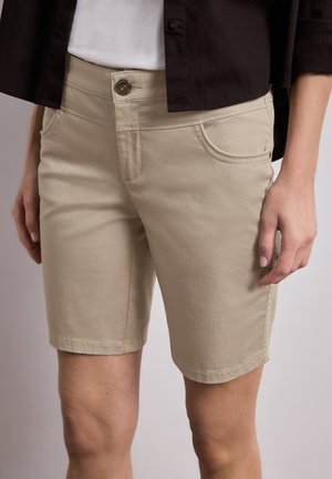 Beige knælange shorts med knap og lynlås, båret med en hvid top og sort åben skjorte, vist fra livet til knæene.