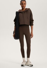 Hoodie corto marrone con mezza zip e design testurizzato, abbinato a leggings marroni. Il modello indossa calze color crema e sneakers color crema con dettagli.