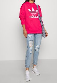 Persona vistiendo una sudadera de Adidas rosa brillante, jeans claros rasgados, zapatillas blancas y llevando un bolso de hombro a rayas azul y blanco.