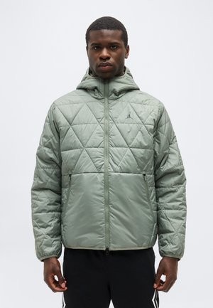 Άνδρας που φοράει ανοιχτό πράσινο καπιτονέ μπουφάν με κουκούλα, στέκεται με το πρόσωπο προς τα εμπρός μπροστά σε απλό φόντο.