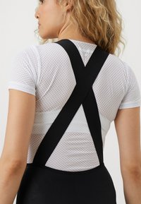 Mulher com cabelo loiro encaracolado usa uma blusa de malha branca de manga curta e suspensórios pretos cruzados nas costas, voltada para a esquerda sobre um fundo branco.