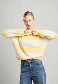 Keltainen ja valkoinen raidoitettu neulepaita, väljä malli, teksturoitu kangas, leveät hihat, yhdistetty sinisiin farkkuihin. Pehmeä, rento muotoilu.