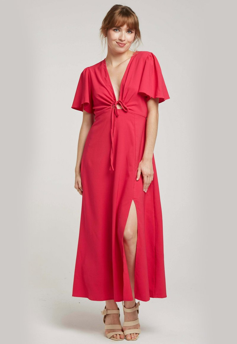 Robe maxi en rose vif, avec un décolleté en V profond, des détails à nouer à la taille, des manches volantées et une fente sur le côté. Texture de tissu lisse.