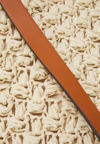 Una cintura in pelle marrone poggia su uno sfondo intrecciato texturizzato di fibre di beige chiaro, creando un contrasto tra superfici lisce e ruvide.