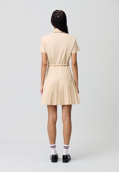 Vestido beige de manga corta con falda plisada, cintura ceñida y cuello. Combinado con zapatos negros y calcetines blancos con rayas rojas y azules.