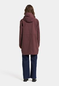 Bordeaux waterdichte jas met capuchon, met een snit tot halverwege de dij, twee zijzakken en een gladde textuur. Draagt met donkere jeans en laarzen.